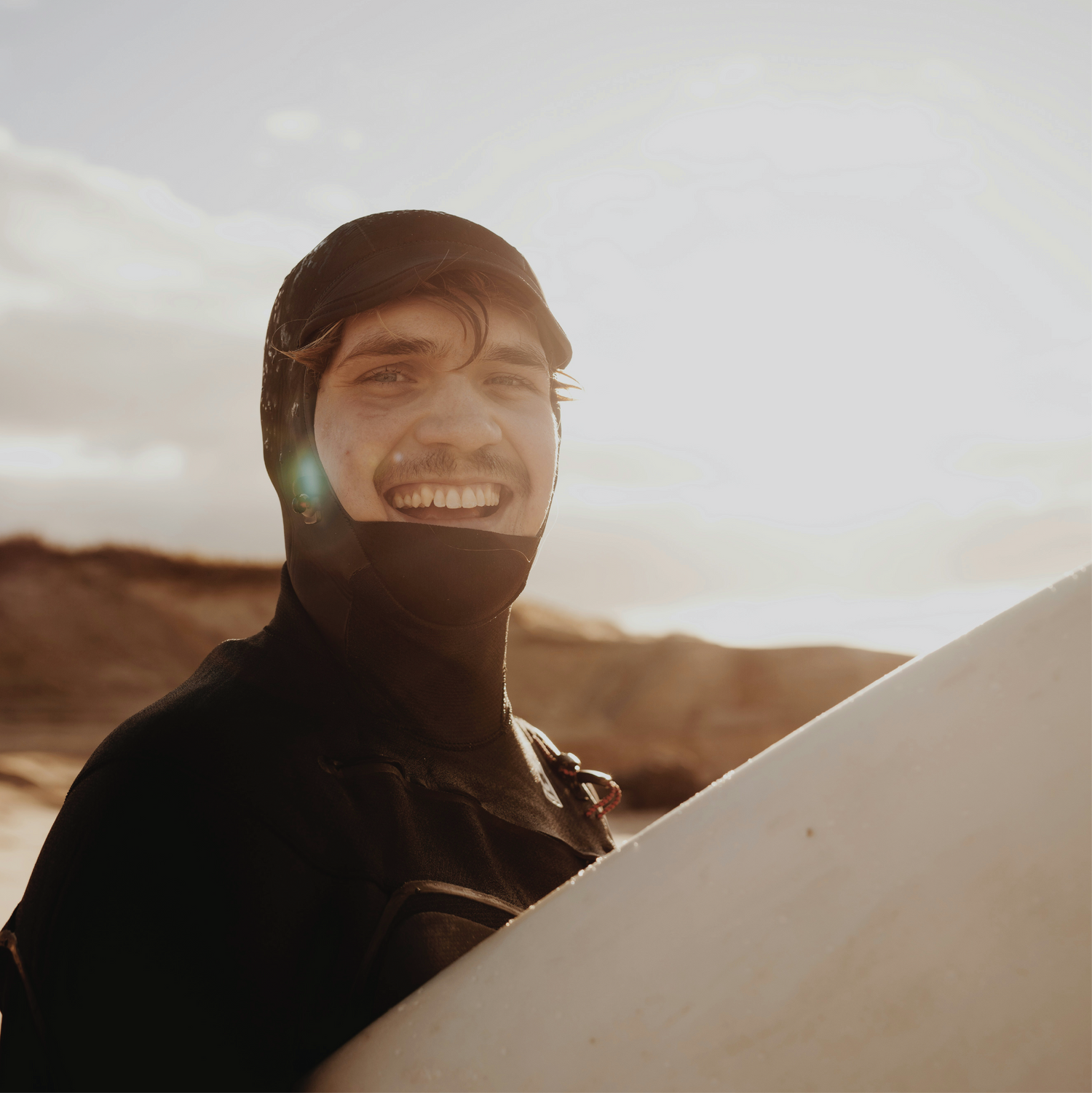 Man smiling about hair loss treatment options, holding surfboard in wetsuit.